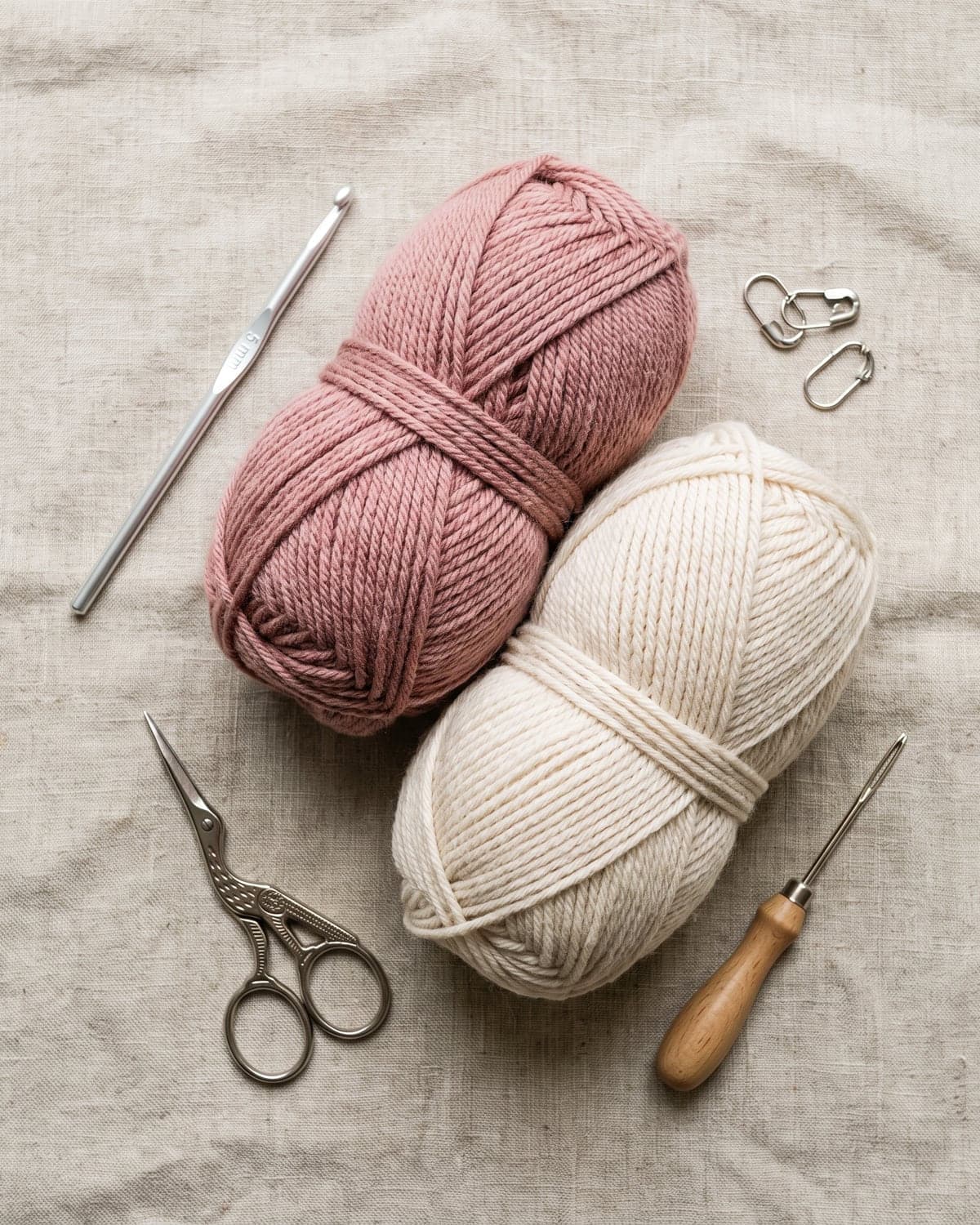 Dusty rose and cream yarn with a crochet hook, scissors and stitch markers on linen