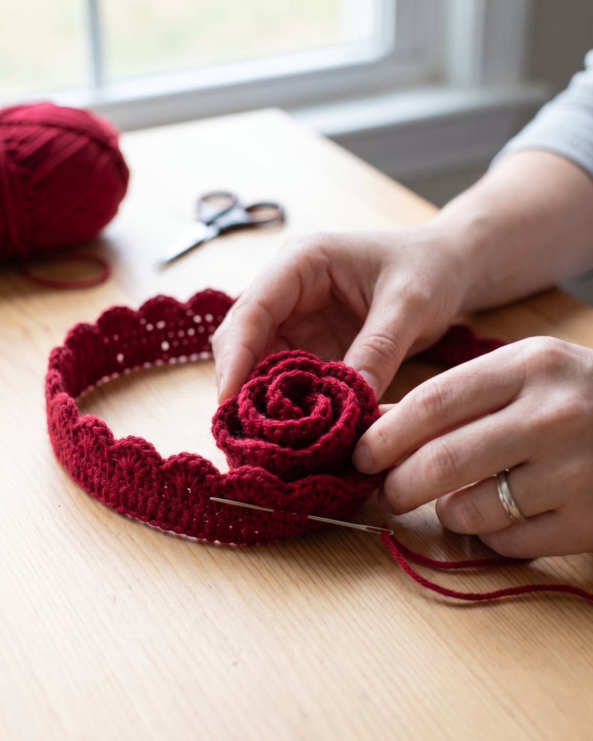 Hands rolling a red scalloped crochet strip into a rose and sewing the base with a needle