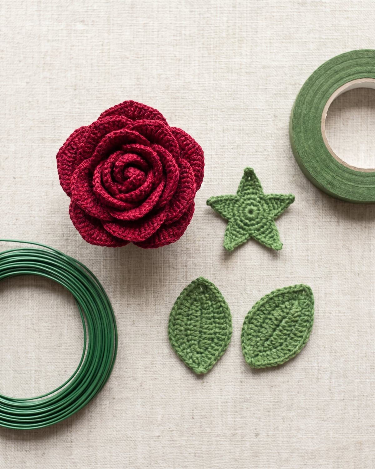 Finished red crochet rose head beside a green star-shaped calyx, two crochet leaves, wire and floral tape