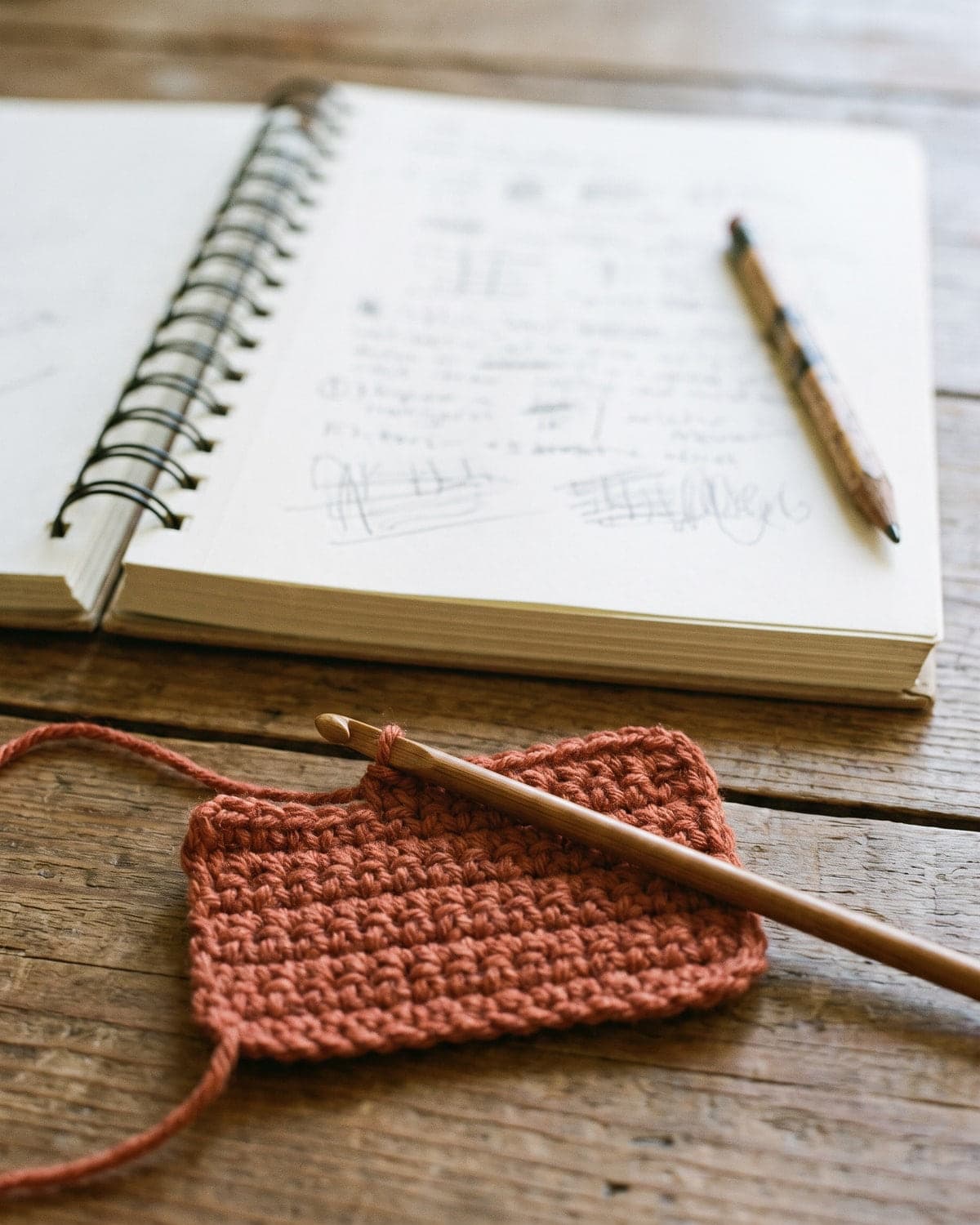 Open spiral notebook with pencil notes beside a terracotta crochet swatch and a wooden hook